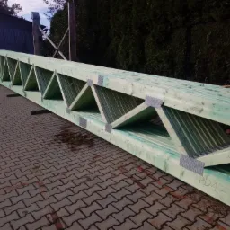Długa, zielona, drewniana więźba dachowa z metalowymi łącznikami, leżąca na brukowanej powierzchni, widoczne oznaczenia 'PD1' i 'S14' na drewnie, tło z zielenią i budynkiem.