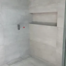 Świeżo wykafelkowana, minimalistyczna kabina prysznicowa z szarą, matową glazurą, odpływem liniowym i wnęką ścienną na kosmetyki, widoczne wystające elementy instalacji wodnej.