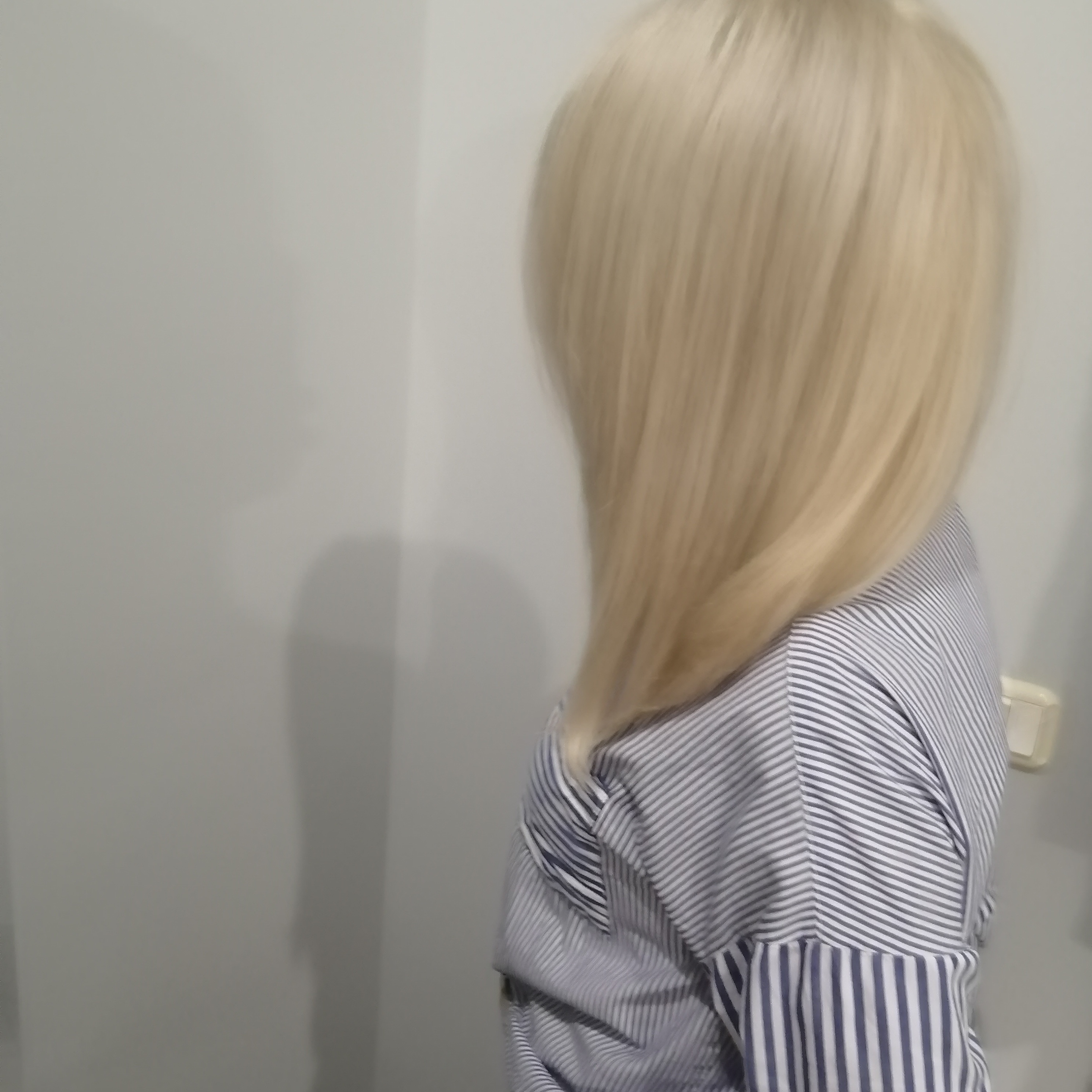 Świeżo ufarbowane, platynowe blond włosy, opadające na ramię w pobliżu ściany, widoczne fragmenty bluzki w biało-szare paski.