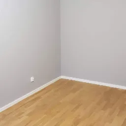Pusty pokój po remoncie: parkiet z jasnego drewna ułożony w jodełkę, szare ściany z białą listwą przypodłogową i sufitową, gniazdko elektryczne na ścianie.