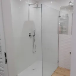 Nowoczesna kabina prysznicowa typu walk-in z chromowaną armaturą i deszczownicą, białe panele ścienne, drewnopodobna podłoga, widok na grzejnik drabinkowy w lustrze.