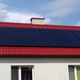 Czarny panel słoneczny na czerwonym dachu domu jednorodzinnego z kremową elewacją, widok z dołu w słoneczny dzień.