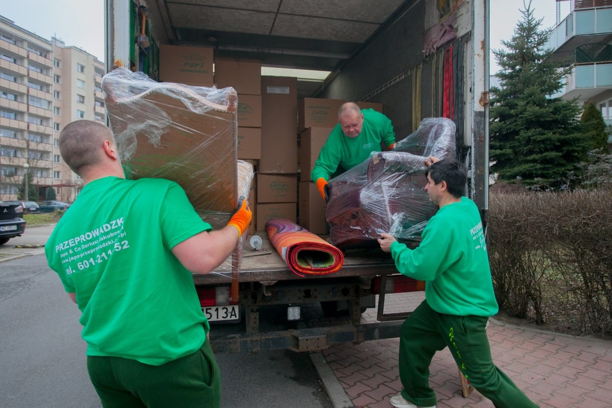 Trzech mężczyzn w zielonych uniformach, z widocznym logo firmy przeprowadzkowej na plecach, ładuje meble owinięte folią i zwinięty dywan do samochodu dostawczego na tle bloku mieszkalnego.