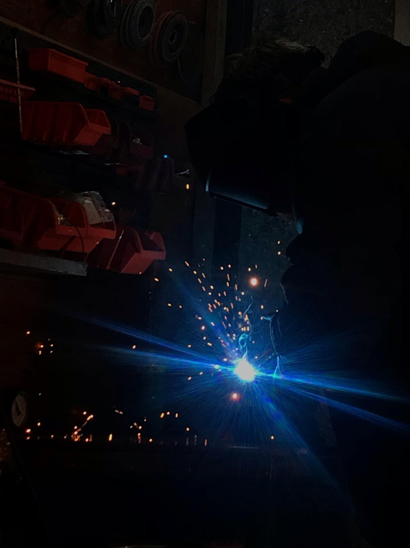 Silueta spawacza w masce ochronnej, pracującego w warsztacie, z intensywnym niebieskim światłem i iskrami podczas procesu łączenia metali. W tle widoczne półki z pojemnikami i oponami.
