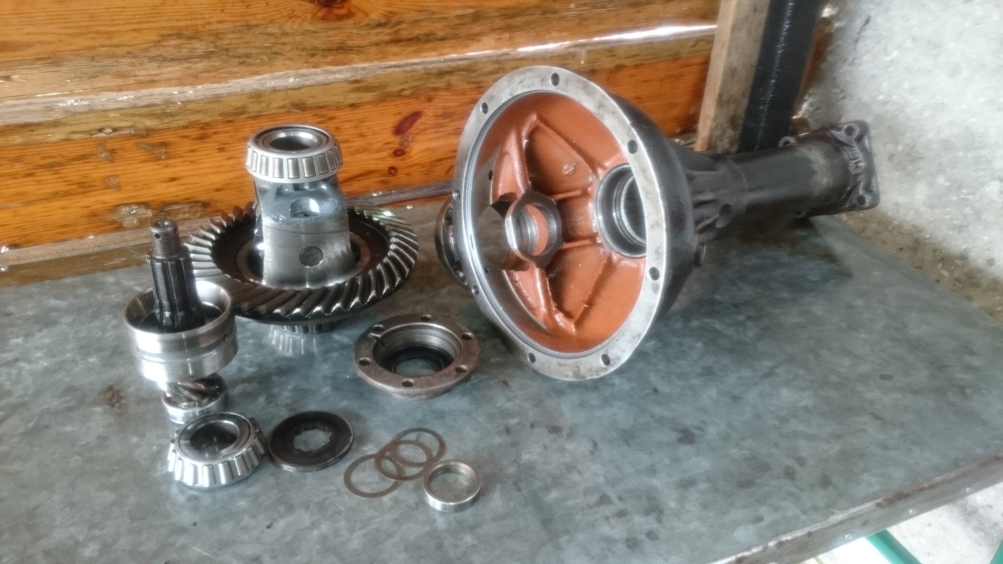 Rozłożony mechanizm różnicowy na metalowym stole, z widocznymi przekładniami zębatymi, łożyskami i obudową z pomarańczowym wnętrzem, na tle drewnianej ściany.