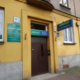Wejście do kancelarii adwokackiej w żółtym budynku z brązowymi drzwiami, szyldami informacyjnymi i numerem budynku 14/I. Nad wejściem balkon z dekoracyjną, metalową balustradą. Na ścianie tablica...