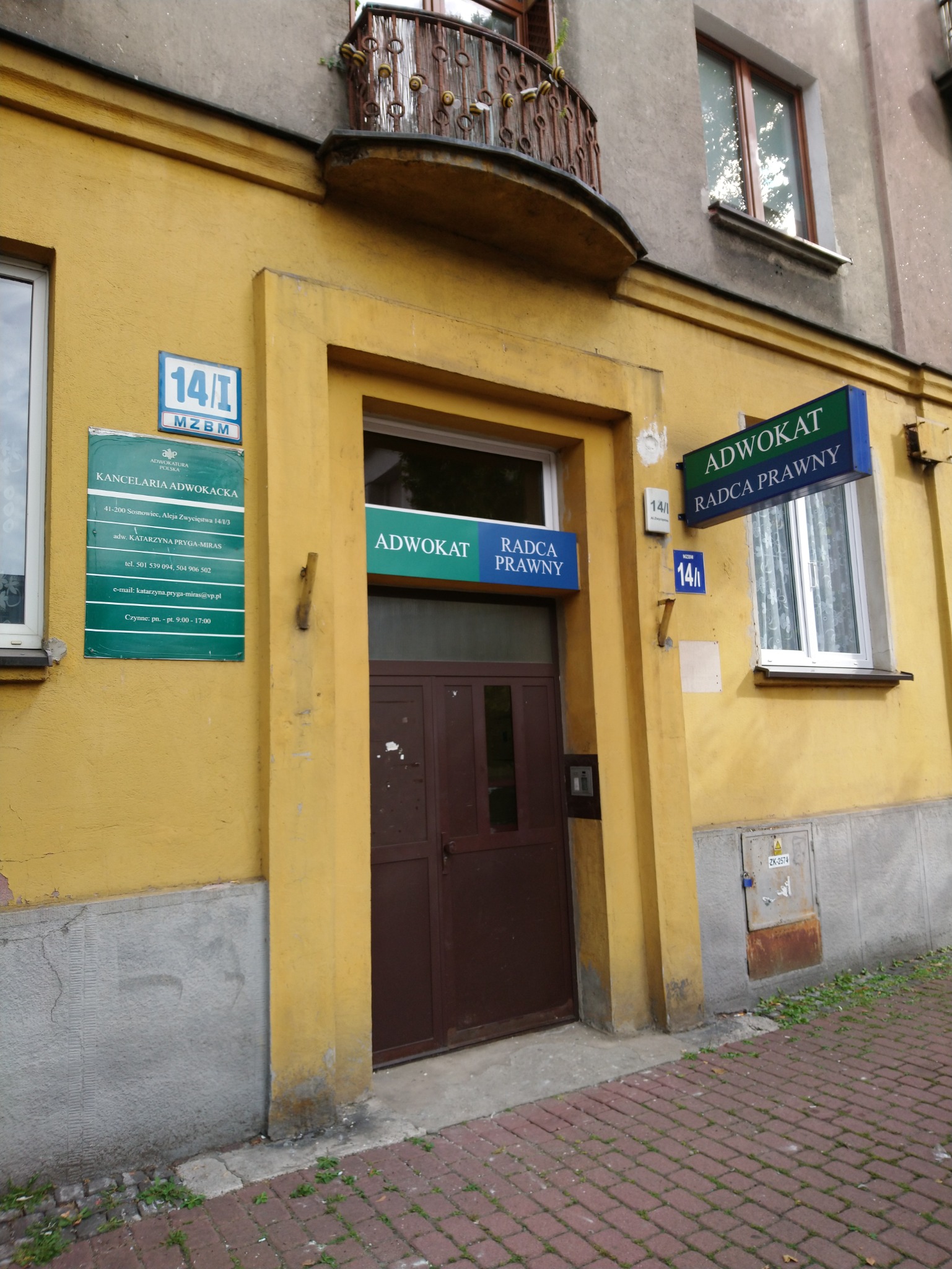 Wejście do kancelarii adwokackiej w żółtym budynku z brązowymi drzwiami, szyldami informacyjnymi i numerem budynku 14/I. Nad wejściem balkon z dekoracyjną, metalową balustradą. Na ścianie tablica...