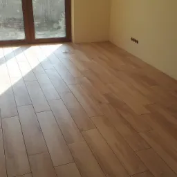 Ułożona podłoga z płytek imitujących drewno w jasnym, pustym pokoju z dużym oknem i czterema gniazdkami elektrycznymi na ścianie.