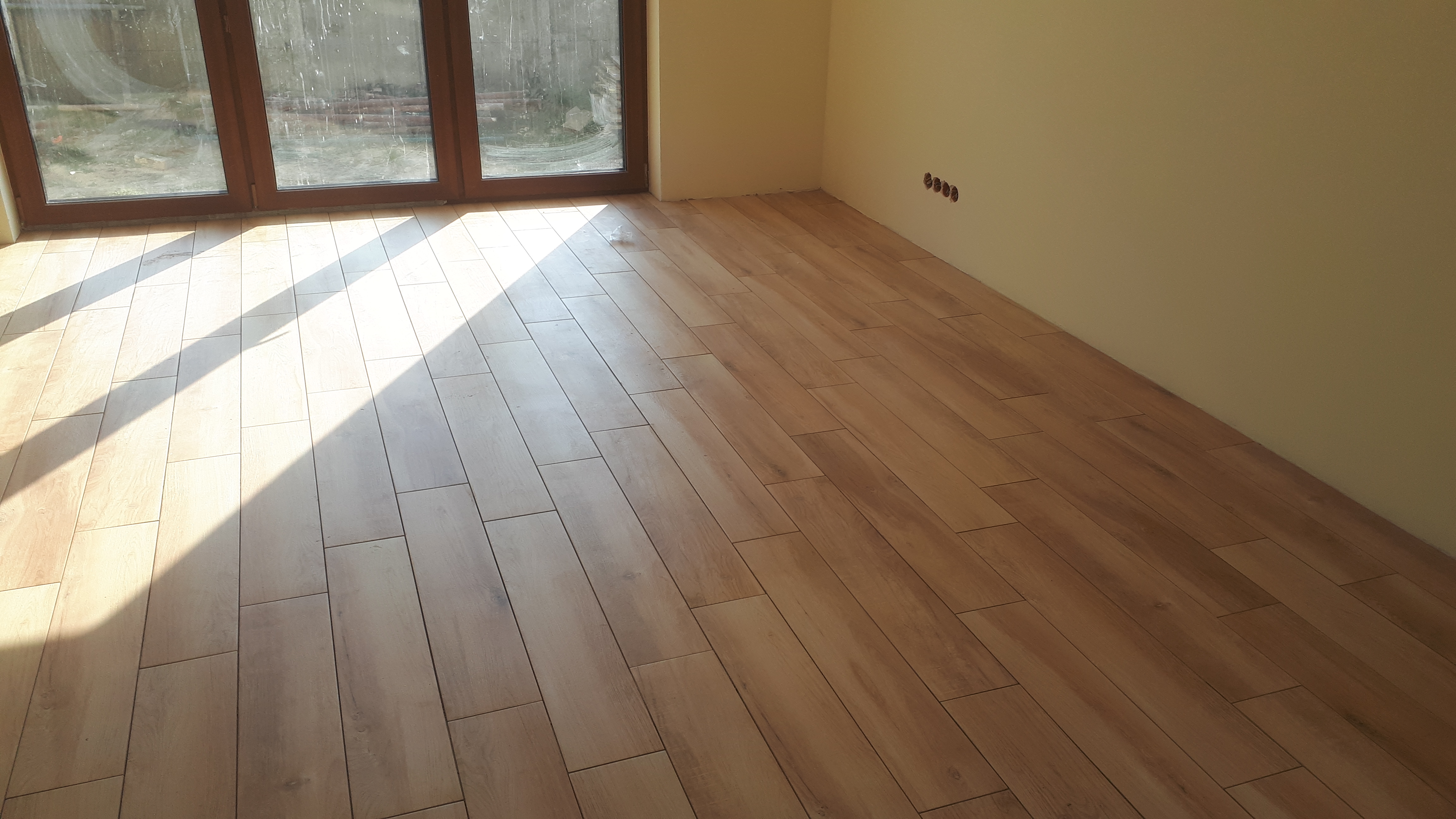 Ułożona podłoga z płytek imitujących drewno w jasnym, pustym pokoju z dużym oknem i czterema gniazdkami elektrycznymi na ścianie.