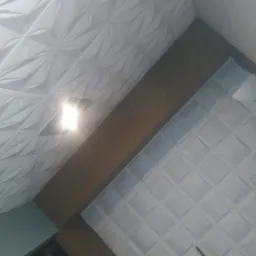 Fragment sufitu z dekoracyjnymi panelami 3D o wzorze geometrycznym, oświetlonym punktowo, z widoczną lampą i gniazdkiem elektrycznym.