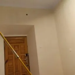 Wnętrze pokoju w trakcie odświeżania, zabezpieczona podłoga folią malarską, widoczny fragment kija teleskopowego do malowania ścian, świeżo pomalowane ściany w odcieniach bieli i beżu, drzwi...