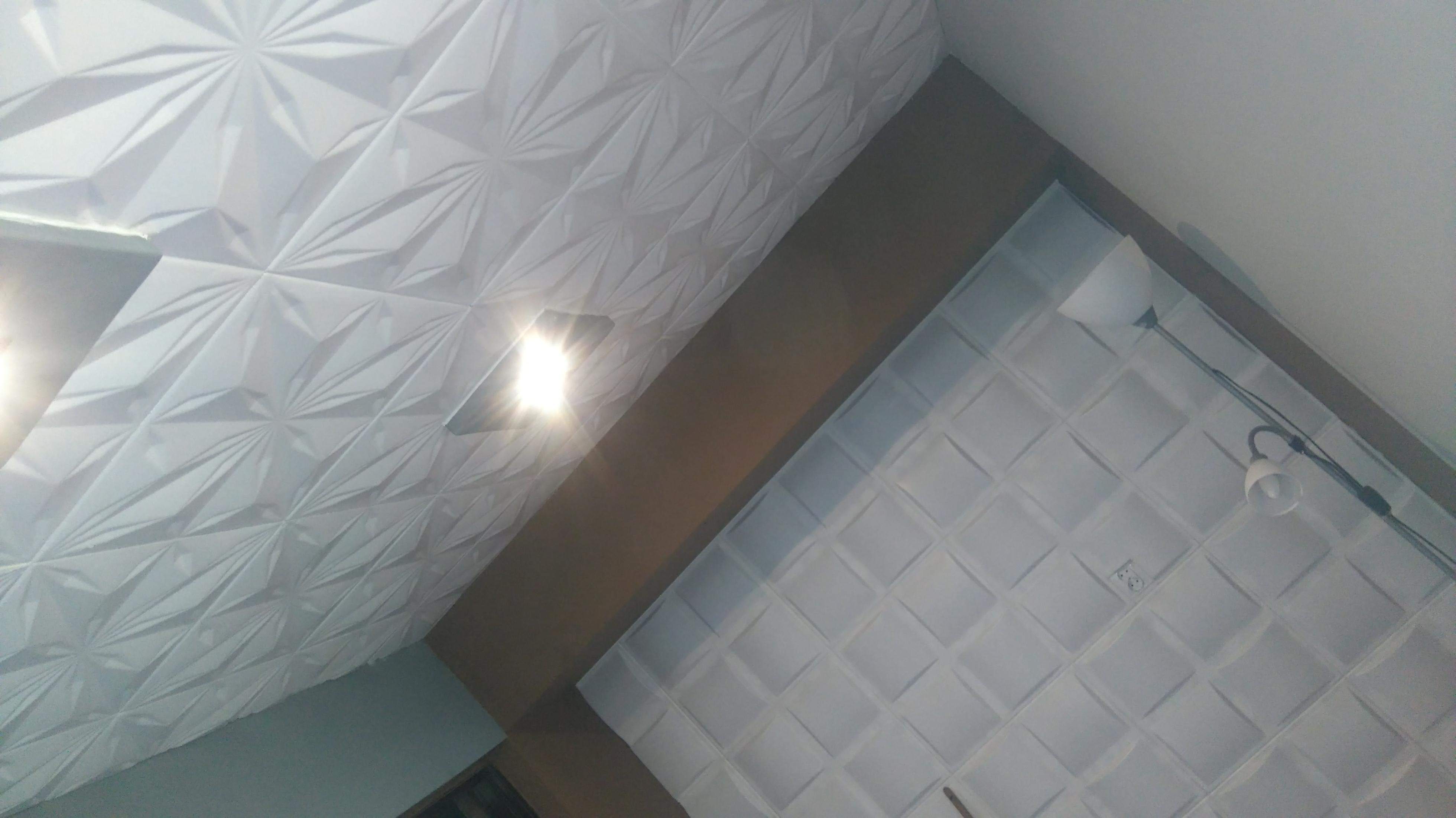 Fragment sufitu z dekoracyjnymi panelami 3D o wzorze geometrycznym, oświetlonym punktowo, z widoczną lampą i gniazdkiem elektrycznym.