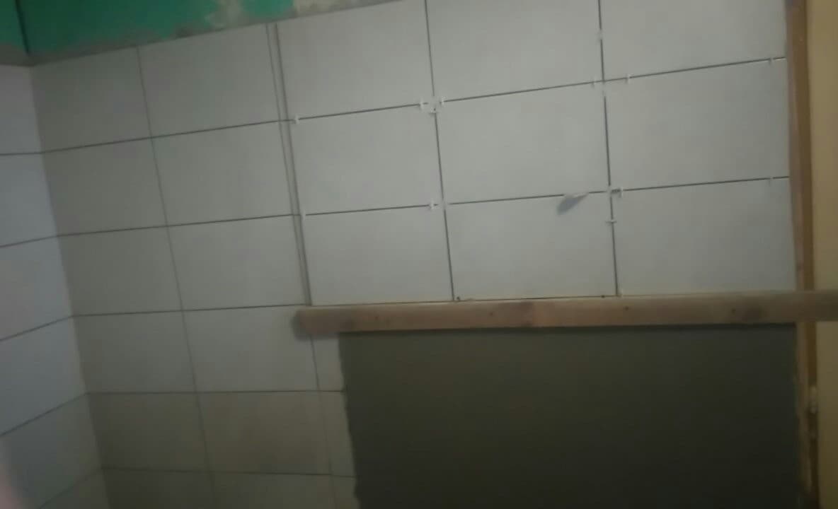 Świeżo ułożone, jasne płytki ceramiczne na ścianie, z widocznymi krzyżykami dystansowymi, częściowo zakryte deską i szarą masą w dolnej części.