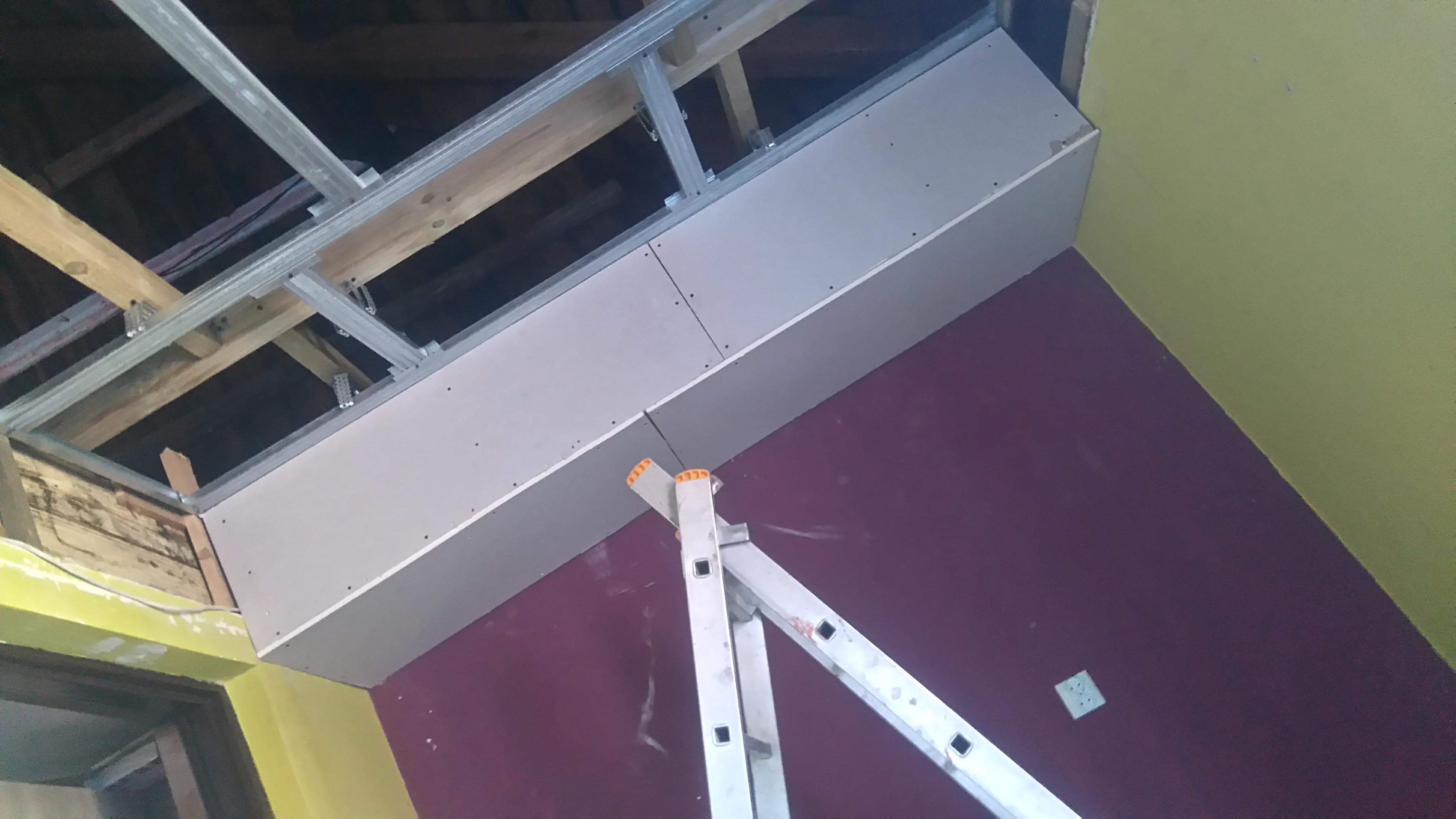 Montaż podwieszanego sufitu z płyt karton gips na metalowej konstrukcji, widoczna drabina, ściany w kolorze żółtym i bordowym.