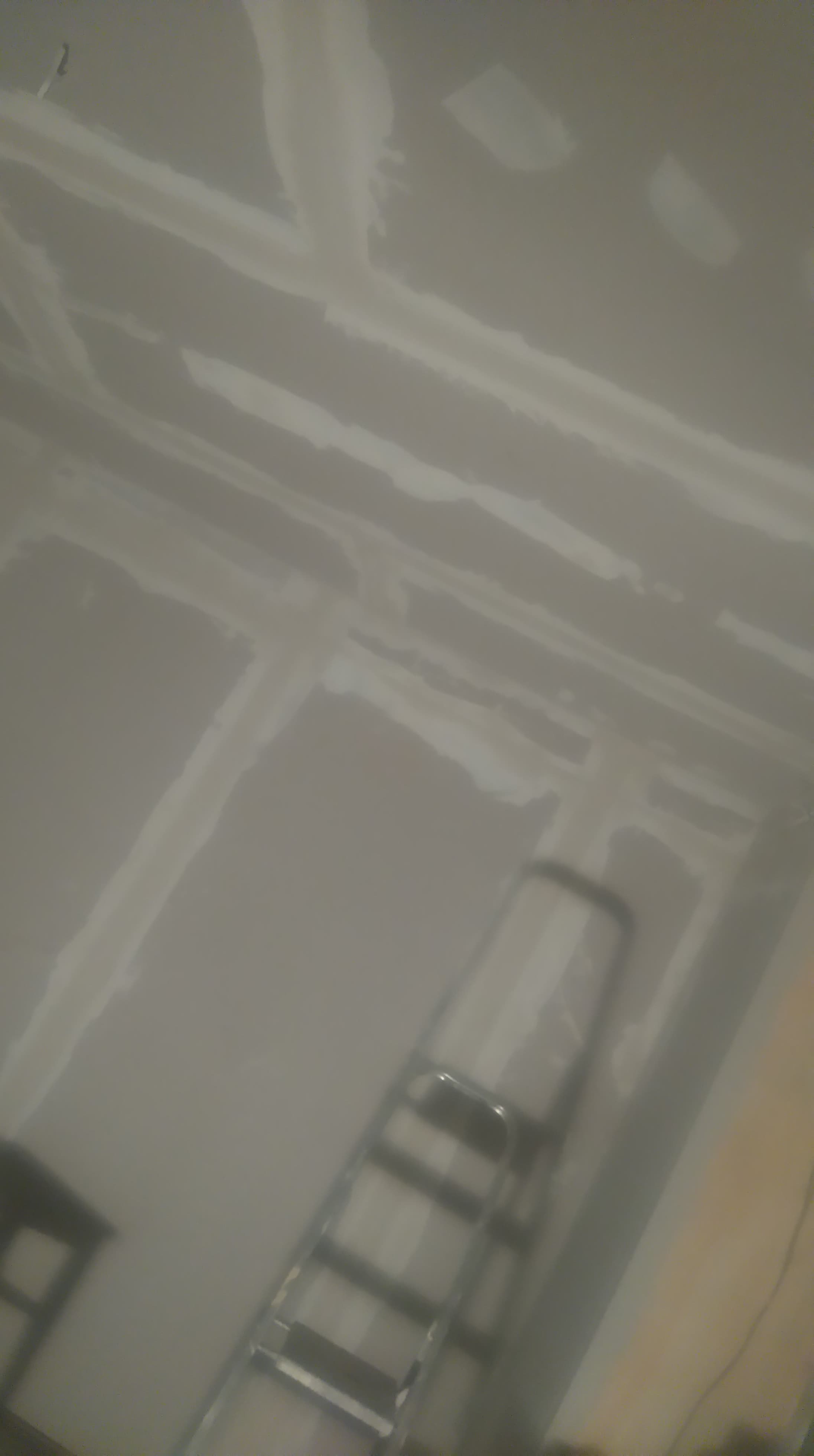 Ściana i sufit po nałożeniu warstwy szpachli, widoczne łączenia płyt kartonowo-gipsowych, drabina w tle, słabe oświetlenie.