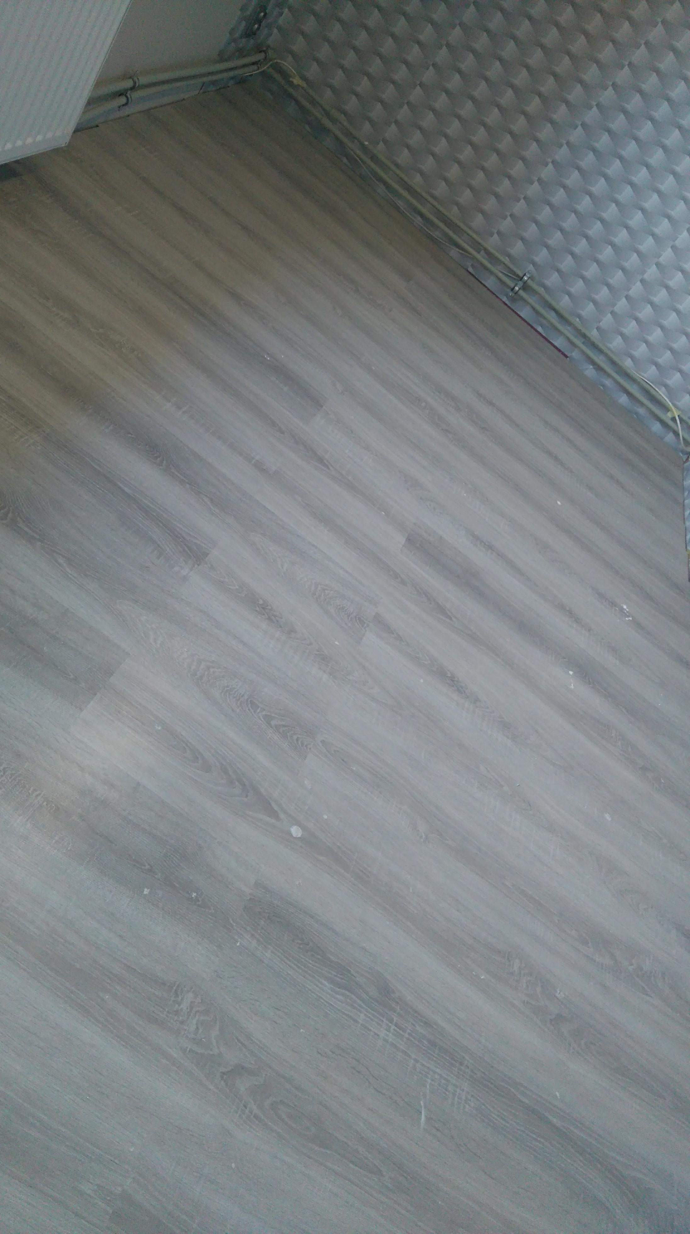 Jasnoszara podłoga z paneli ułożona ukośnie, widoczne rury przy suficie i fragment sufitu z fakturą kostki.