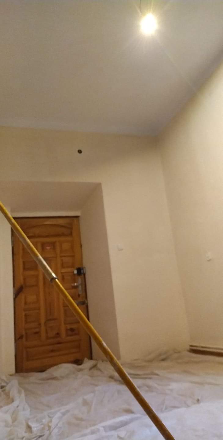 Wnętrze pokoju w trakcie odświeżania, zabezpieczona podłoga folią malarską, widoczny fragment kija teleskopowego do malowania ścian, świeżo pomalowane ściany w odcieniach bieli i beżu, drzwi...