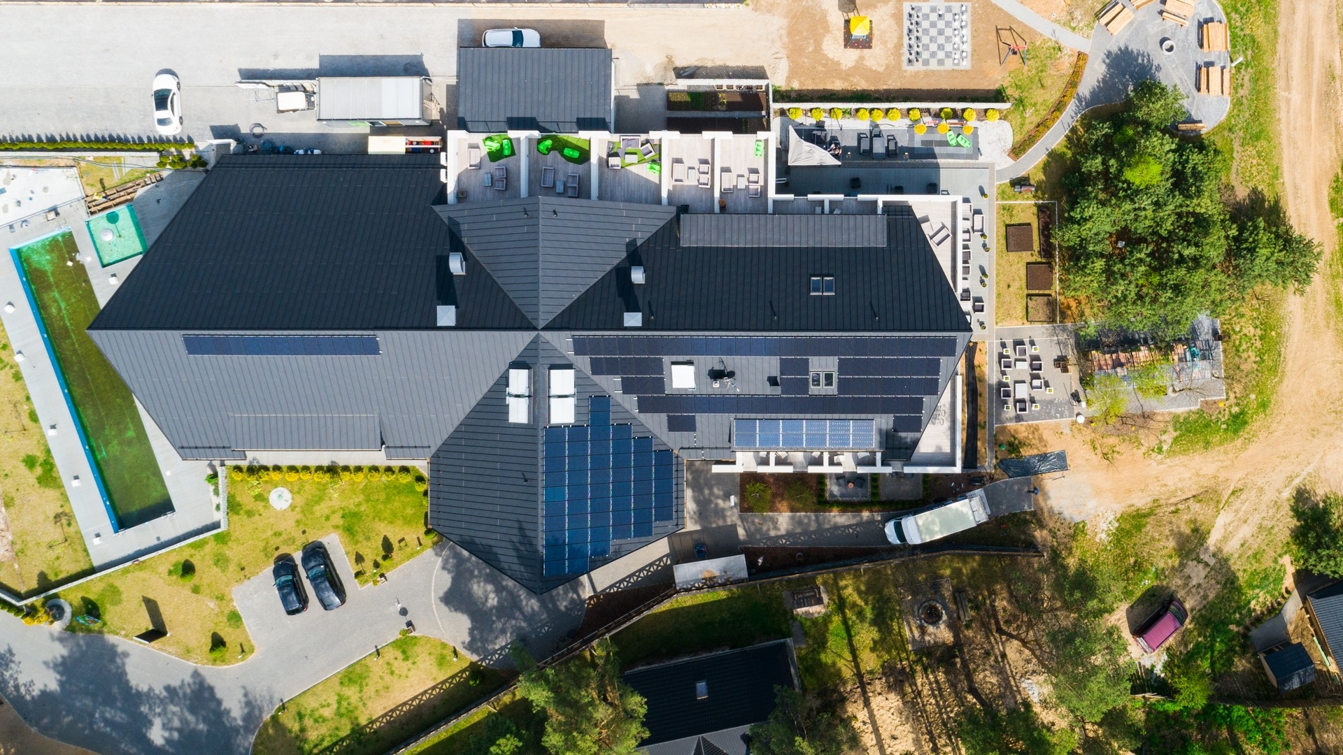 Widok z lotu ptaka na duży dom z ciemnym dachem, na którym zamontowano panele słoneczne. Obok domu znajduje się basen, parking dla samochodów, taras z meblami ogrodowymi oraz plac zabaw.