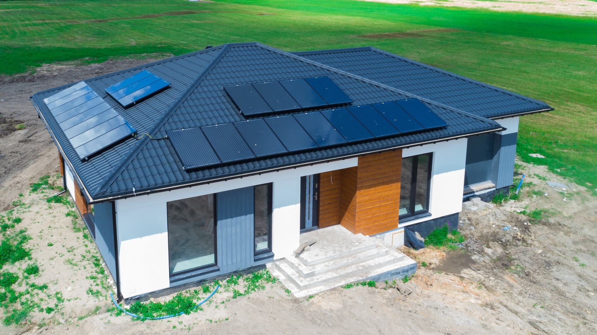 Nowoczesny dom jednorodzinny z ciemną dachówką i panelami słonecznymi, widok z góry, teren wokół domu w trakcie zagospodarowania.