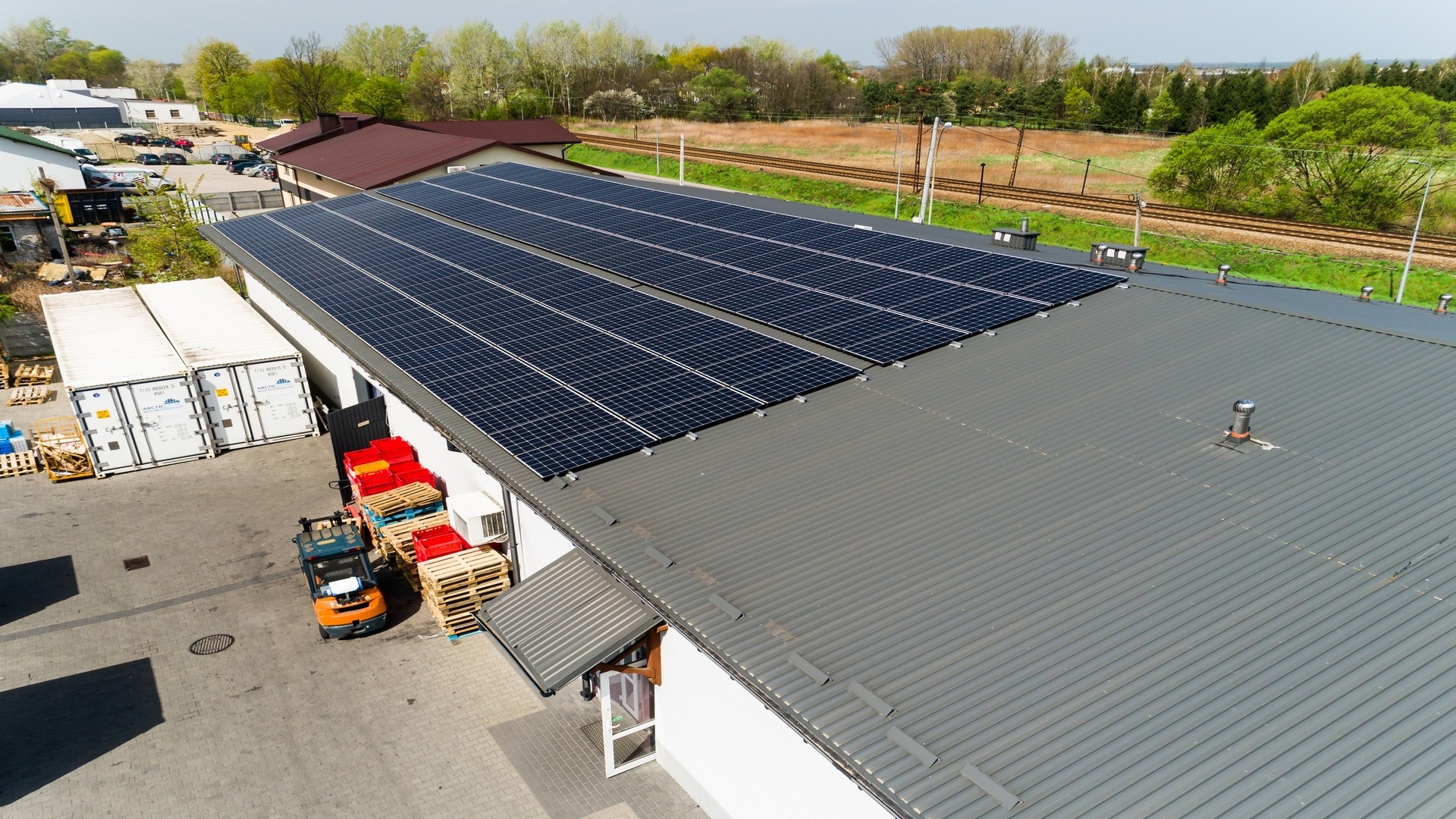 Widok z góry na budynek z szarym dachem, na którym zamontowane są panele fotowoltaiczne. Obok budynku stoją białe kontenery, palety i wózek widłowy. W tle widoczne drzewa i tor kolejowy.