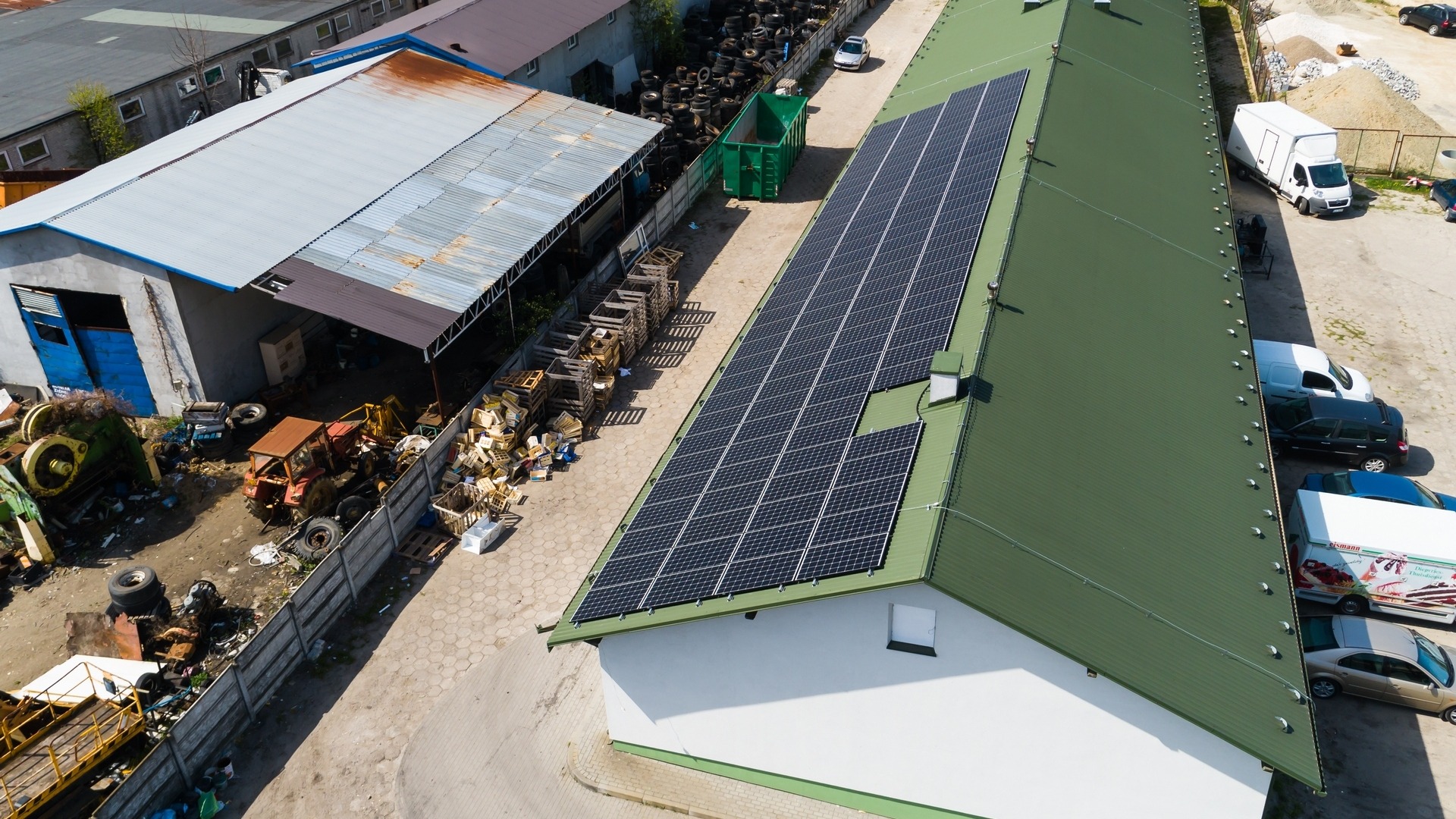 Widok z góry na budynek z panelami słonecznymi na dachu, obok składowisko złomu i opon, oraz zaparkowane samochody dostawcze.