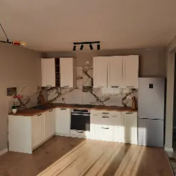 Jasna kuchnia w stylu nowoczesnym z białymi szafkami, drewnianymi blatami i marmurowymi płytkami na ścianie, oświetlona lampami sufitowymi i wiszącą lampą z żarówką edisona.