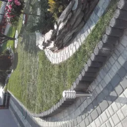 Ułożona szara kostka brukowa z obrzeżami z betonowych palisad oddzielająca trawnik i rabatę skalną z roślinami, oświetlenie ogrodowe w tle.