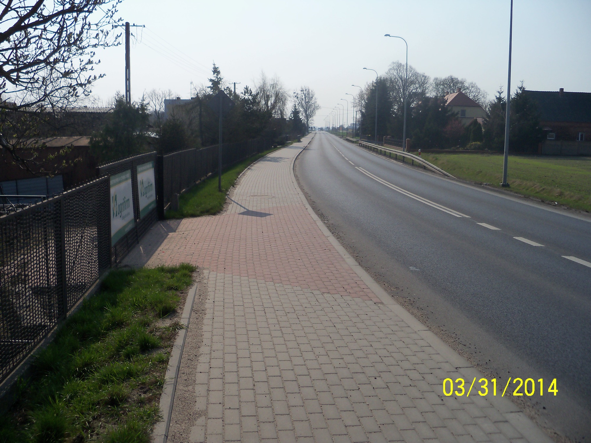 Długa, prosta droga z chodnikiem wyłożonym kostką brukową w odcieniach szarości i czerwieni, widok z perspektywy pieszego, w tle linia drzew i zabudowania.