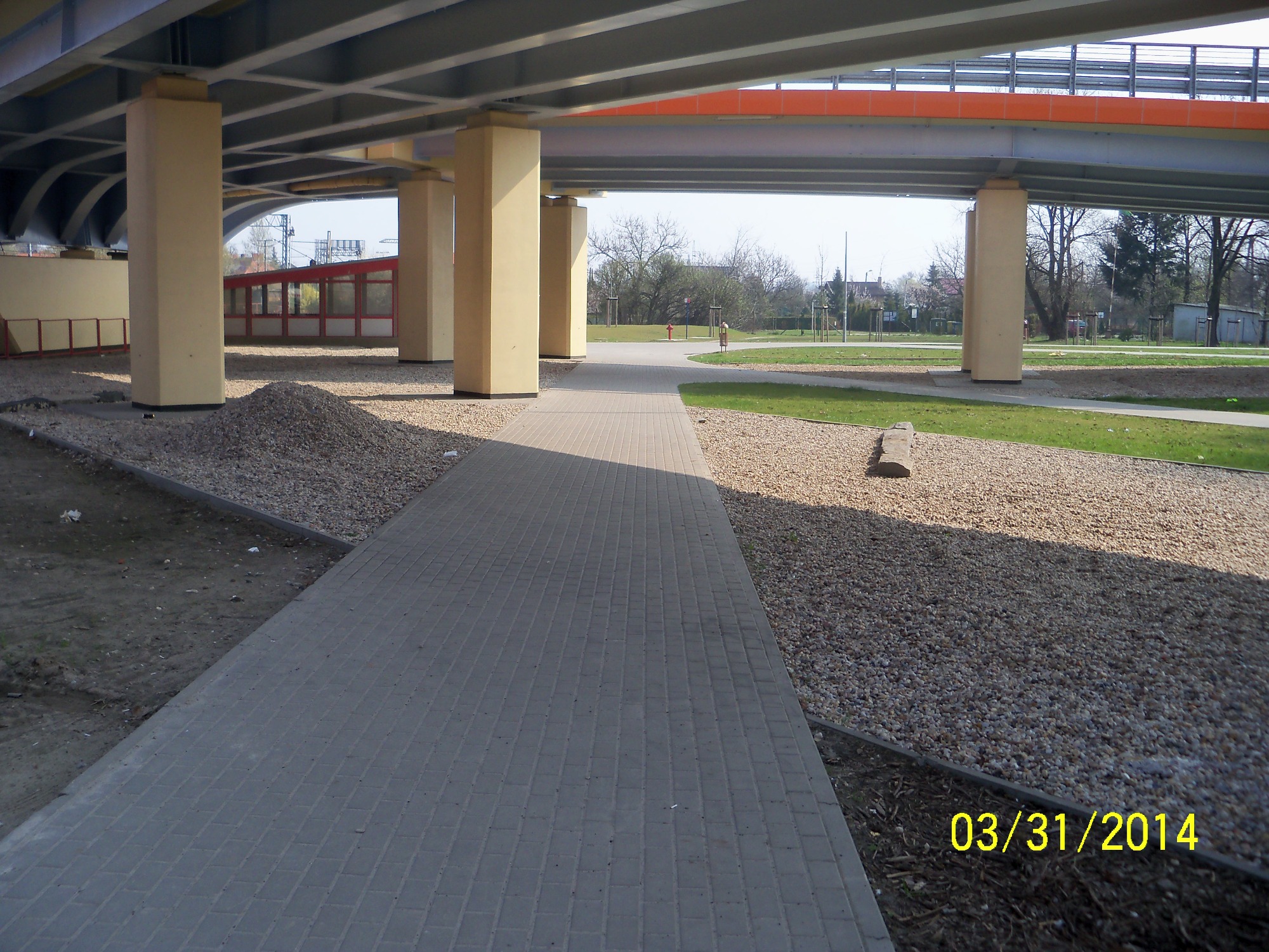 Szara ścieżka z kostki brukowej prowadząca pod wiaduktem, otoczona żwirem i trawnikiem, z widocznym stosem żwiru po lewej stronie.