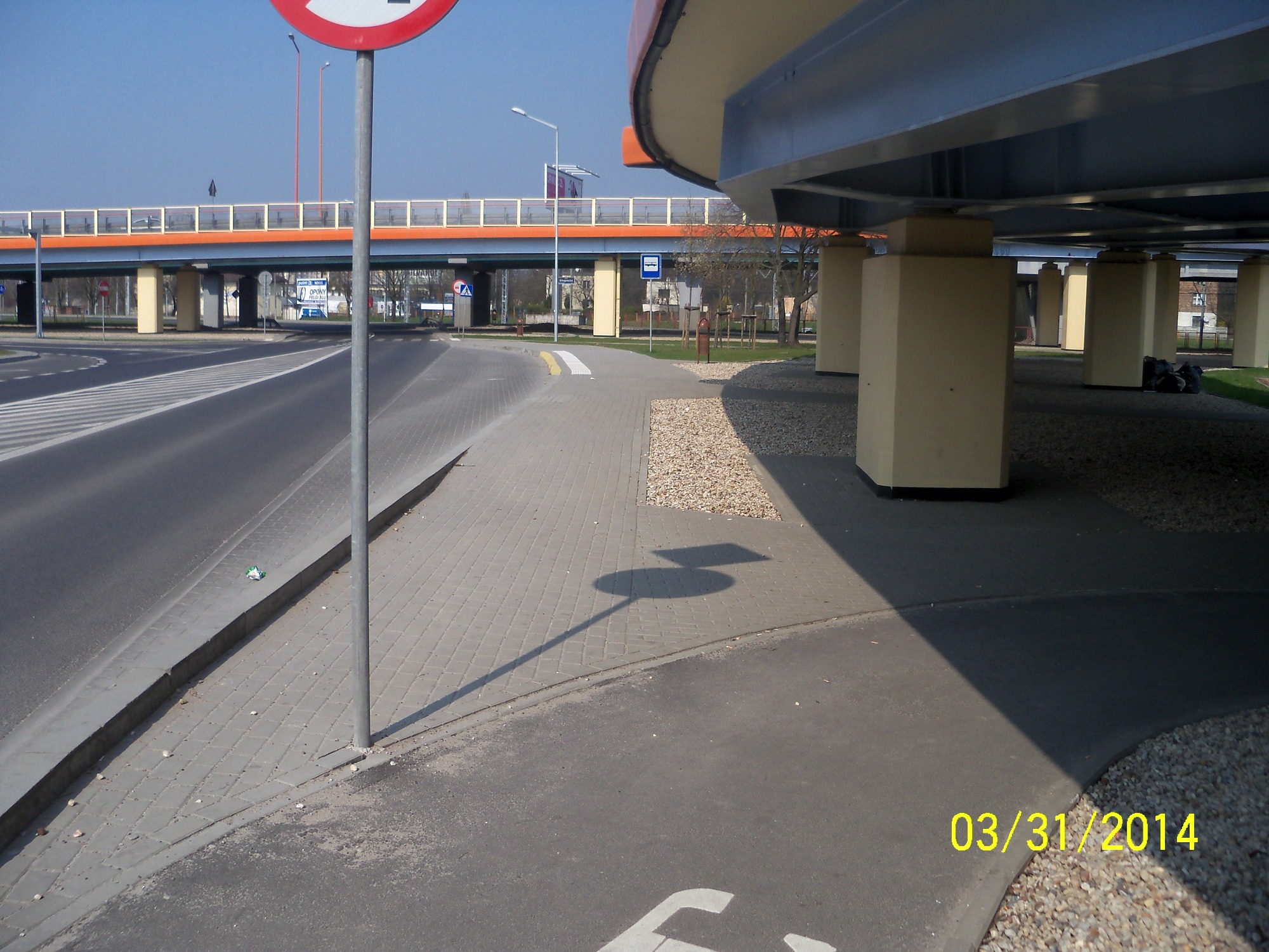 Szara kostka brukowa na chodniku wzdłuż jezdni, cień znaku drogowego, w tle wiadukt z pomarańczowymi barierkami i filary podtrzymujące konstrukcję.
