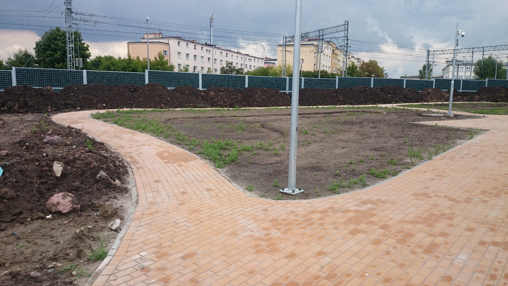 Ścieżka z jasnej kostki brukowej rozwidlająca się na dwie strony, obok świeżo usypana ziemia, w tle budynki mieszkalne i słupy energetyczne.