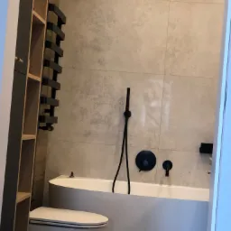 Nowoczesna łazienka z beżowymi płytkami, białą wanną i czarną armaturą, widok zza drzwi.