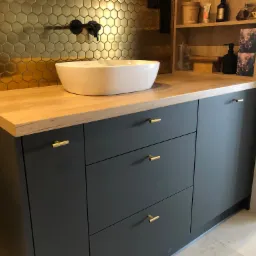 Umywalka nablatowa w kształcie misy na drewnianym blacie, pod nią szafka z szufladami w kolorze grafitowym z złotymi uchwytami, w tle ściana wyłożona złotą mozaiką w kształcie plastra miodu...