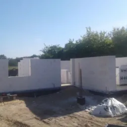 Wznoszone ściany domu z bloczków betonu komórkowego H+H, widoczne zbrojenia i fundamenty, w tle zieleń drzew.