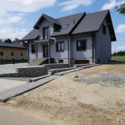 Nowoczesny dom jednorodzinny z szarą elewacją, wykończony kamieniem dekoracyjnym u podstawy, z brukowanym podjazdem i placem przed wejściem, w trakcie prac wykończeniowych na działce.