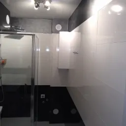 Nowoczesna łazienka z kabiną prysznicową, białymi płytkami ściennymi i czarnymi akcentami, oświetlona lampą sufitową z kilkoma reflektorami.