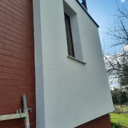 Fragment elewacji budynku z oknem, łączący ceglaną ścianę z otynkowaną, pomalowaną na biało, widoczne rusztowanie.