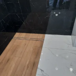 Nowoczesna łazienka z kontrastującym połączeniem płytek: drewnopodobnych, białych marmurkowych i czarnych marmurkowych, z widocznym odpływem liniowym i złotą baterią podtynkową.