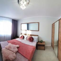 OREST - Jasna sypialnia z drewnianym łóżkiem, lustrem w ramie, szafą z lustrzanymi drzwiami i okrągłymi poduszkami dekoracyjnymi na łóżku.