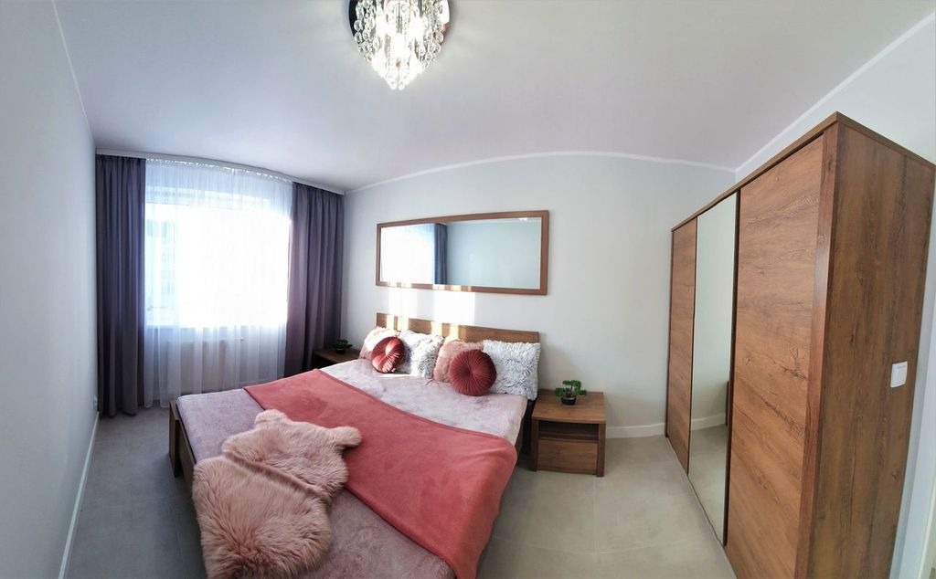 Jasna sypialnia z drewnianym łóżkiem, lustrem w ramie, szafą z lustrzanymi drzwiami i okrągłymi poduszkami dekoracyjnymi na łóżku.