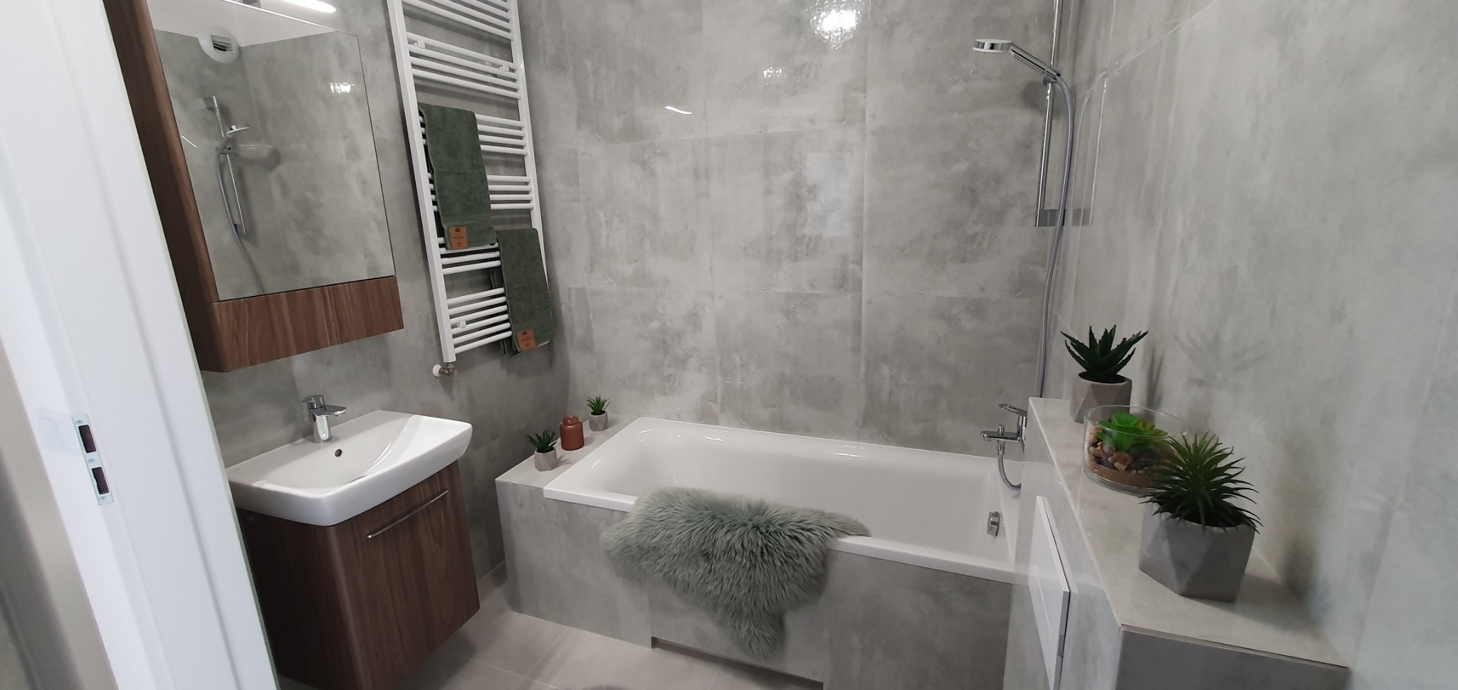 Nowoczesna łazienka z wanną obudowaną szarymi płytkami imitującymi beton, wiszącą szafką z umywalką w kolorze drewna oraz grzejnikiem drabinkowym z zielonymi ręcznikami.