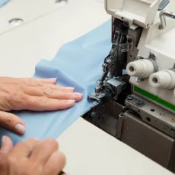 Praca na owerloku: ręce przesuwają błękitny materiał pod stopką maszyny, widoczne detale mechanizmu szyjącego.