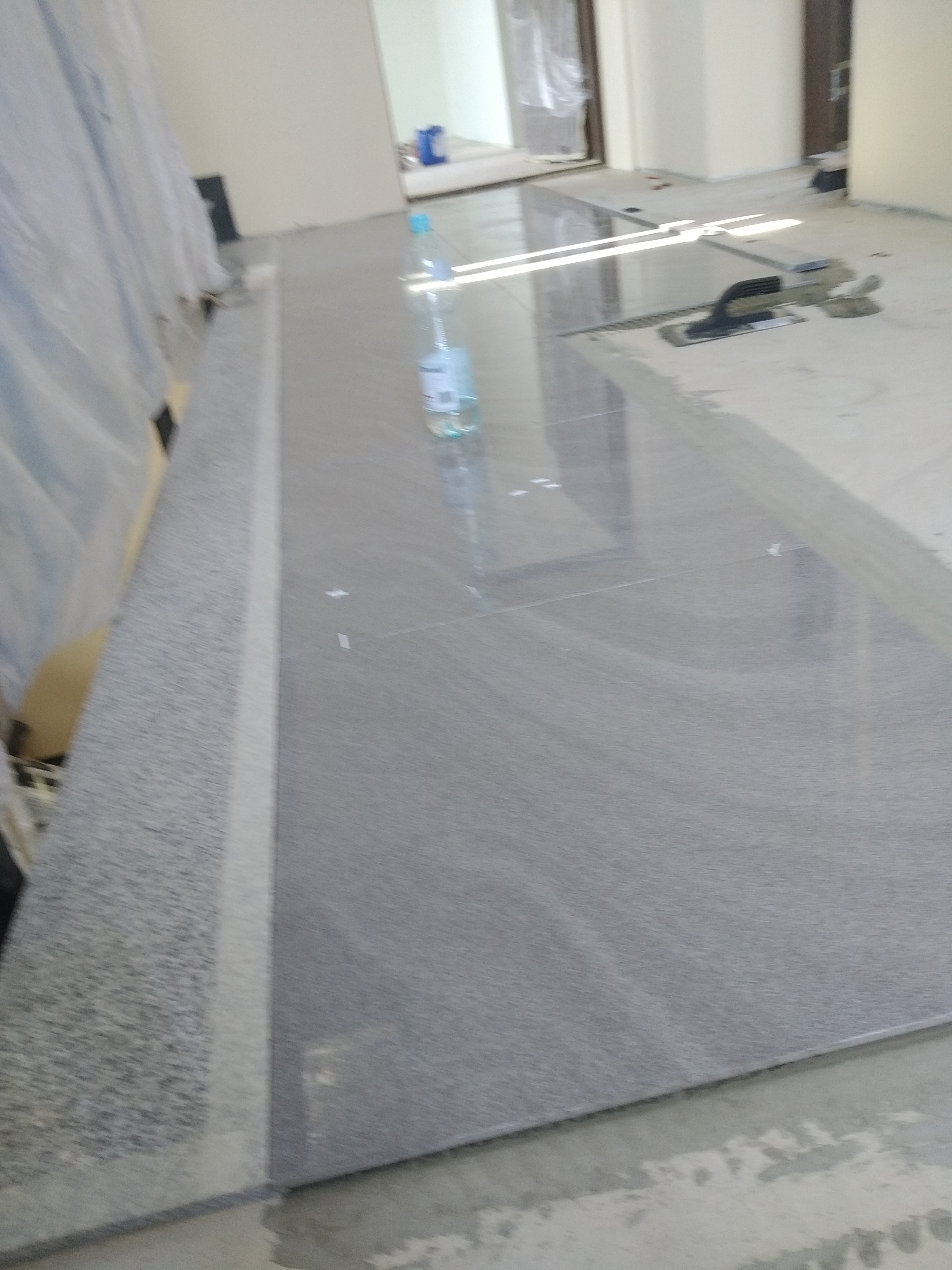 Szare, matowe płytki podłogowe ułożone w pomieszczeniu w trakcie prac wykończeniowych, widoczna butelka wody i narzędzia na powierzchni płytek.