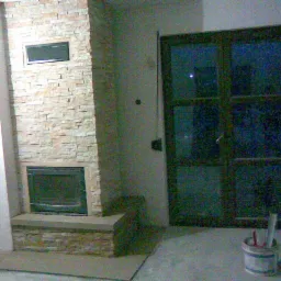 Murowany kominek z jasnego kamienia z czarnym wkładem kominkowym, widoczny w trakcie prac wykończeniowych, obok ciemne drzwi balkonowe i wiadro z narzędziami.