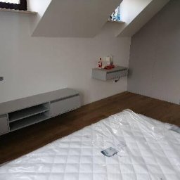 Flormeb - Pokój na poddaszu z drewnianą podłogą, szarą szafką RTV, półką wiszącą i materacem zabezpieczonym folią, przygotowany do dalszego wykończenia.