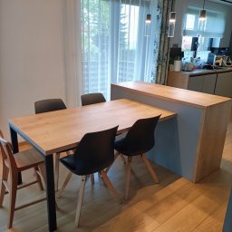 Flormeb - Drewniany stół z czarnymi nogami otoczony czarnymi i drewnianymi krzesłami w nowoczesnej jadalni z parkietem i wyspą kuchenną w tle.