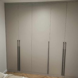 Flormeb - Czterodrzwiowa szafa w odcieniach szarości z minimalistycznymi, długimi, czarnymi uchwytami, ustawiona na parkiecie w jodełkę, z widoczną lampą ścienną i fragmentem łóżka z pościelą.