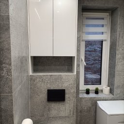 Flormeb - Biała szafka łazienkowa na wymiar z połyskiem, zamontowana nad toaletą w nowoczesnej łazience wykończonej szarą płytką imitującą beton. W tle okno z roletą dzień-noc i dekoracjami.
