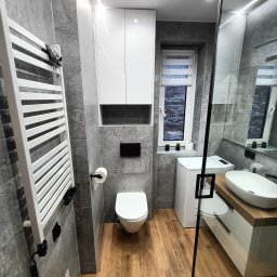 Flormeb - Nowoczesna łazienka z szarymi płytkami, drewnianą podłogą i białymi meblami, w tym szafką nad toaletą, grzejnikiem drabinkowym i umywalką z drewnianym blatem, oświetlona listwami LED.