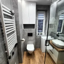 Nowoczesna łazienka z szarymi płytkami, drewnianą podłogą i białymi meblami, w tym szafką nad toaletą, grzejnikiem drabinkowym i umywalką z drewnianym blatem, oświetlona listwami LED.